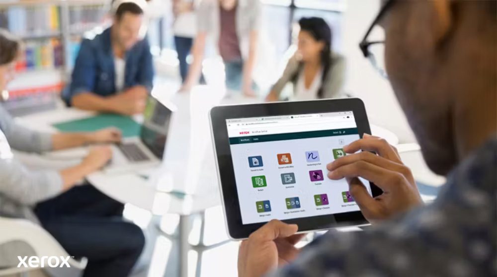 Xerox® Workflow Central Platform: Intuïtieve mobiele toegang Een persoon houdt een tablet vast waarop de interface van Xerox Workflow Central met diverse app-pictogrammen wordt weergegeven, terwijl anderen tegen een wazige achtergrond werken, waardoor de mobiele toegang tot workflowoplossingen wordt benadrukt.