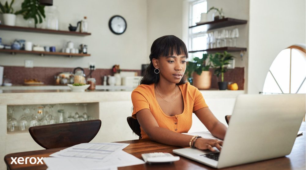 Xerox® DocuShare® Go: thuiswerken mogelijk maken Een persoon zit aan een keukentafel en werkt geconcentreerd op een laptop, met papieren en een rekenmachine in de buurt. Dit illustreert de flexibiliteit van thuiswerken met efficiënt documentbeheer.