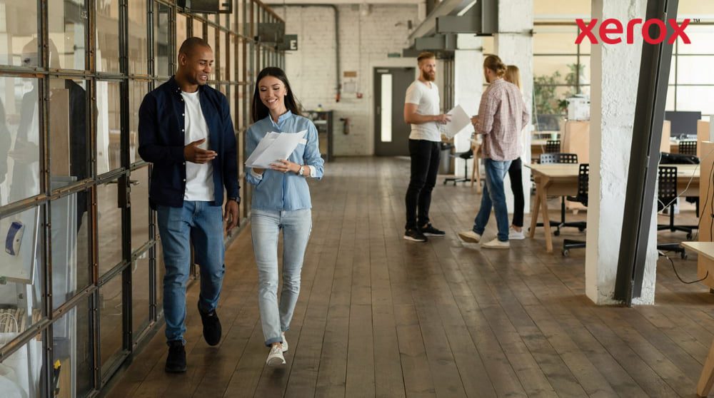 Twee collega's lopen door een gang in een modern, open kantoor. Ze hebben documenten in hun handen en lachen. Op de achtergrond zijn andere collega's zichtbaar, wat symbool staat voor een dynamische en op samenwerking gerichte werkplek.
