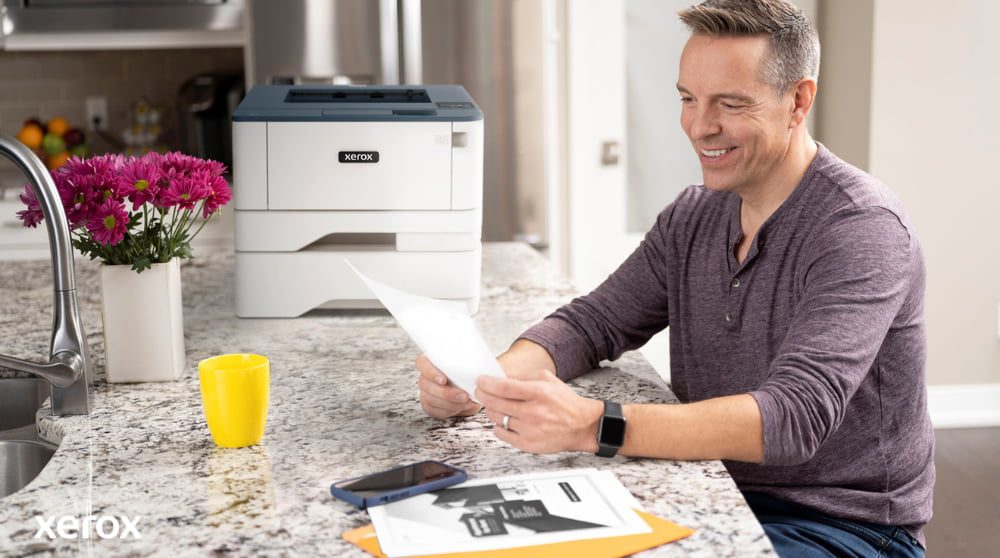"Xerox® Print- en scanervaring: Moeiteloze documentverwerking " Een persoon zit glimlachend aan een keukentafel een document te bekijken. Er staat een compacte Xerox-printer en een felgele mok in de buurt. Dit suggereert dat het printen en verwerken van documenten probleemloos verloopt, thuis of op kantoor.
