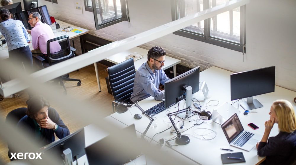 Xerox® Print and Scan Experience: de productiviteit op kantoor verhogen Een bovenaanzicht van een open kantoorruimte met meerdere mensen die achter hun bureau werken en computers gebruiken. De scène straalt een productieve en collaboratieve werkomgeving uit.