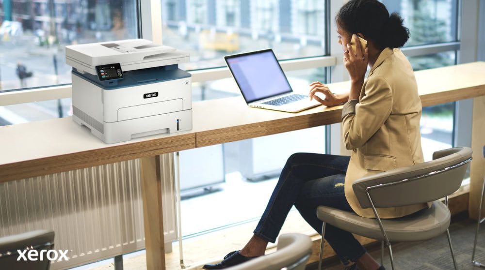 Expérience d'impression et de numérisation Xerox® : pratique et connectée Une personne est assise à une table haute près d'une grande fenêtre, parlant au téléphone tout en travaillant sur un ordinateur portable. Une imprimante multifonction Xerox B235DNI est judicieusement placée sur la table à côté d'elle, suggérant une utilisation simple dans un espace de travail moderne.