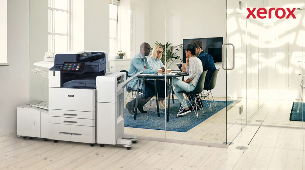 Een moderne Xerox AltaLink B8245 multifunctionele printer staat op de voorgrond van een helder kantoor. Op de achtergrond is een groep mensen aan een tafel in gesprek.