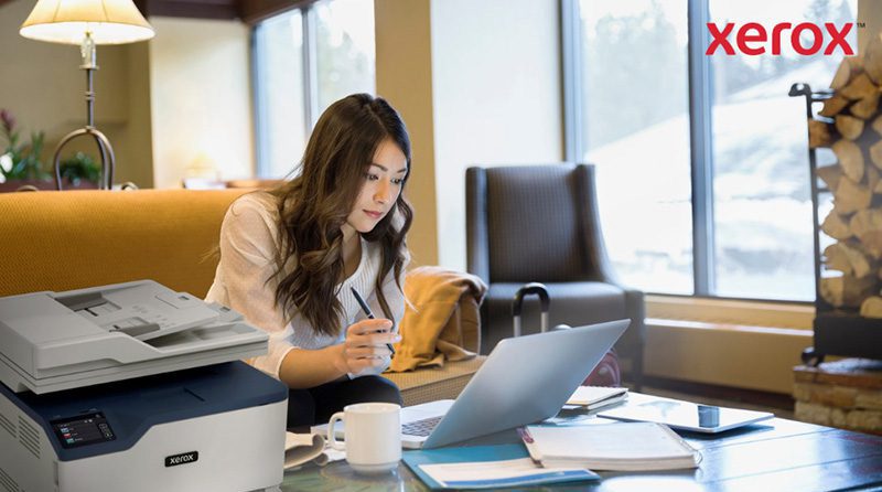 Een vrouw werkt met een laptop in een comfortabele kantoor- of loungeomgeving. Op de voorgrond is een compacte Xerox-printer zichtbaar, wat duidt op een naadloze integratie van printen met digitale workflows.