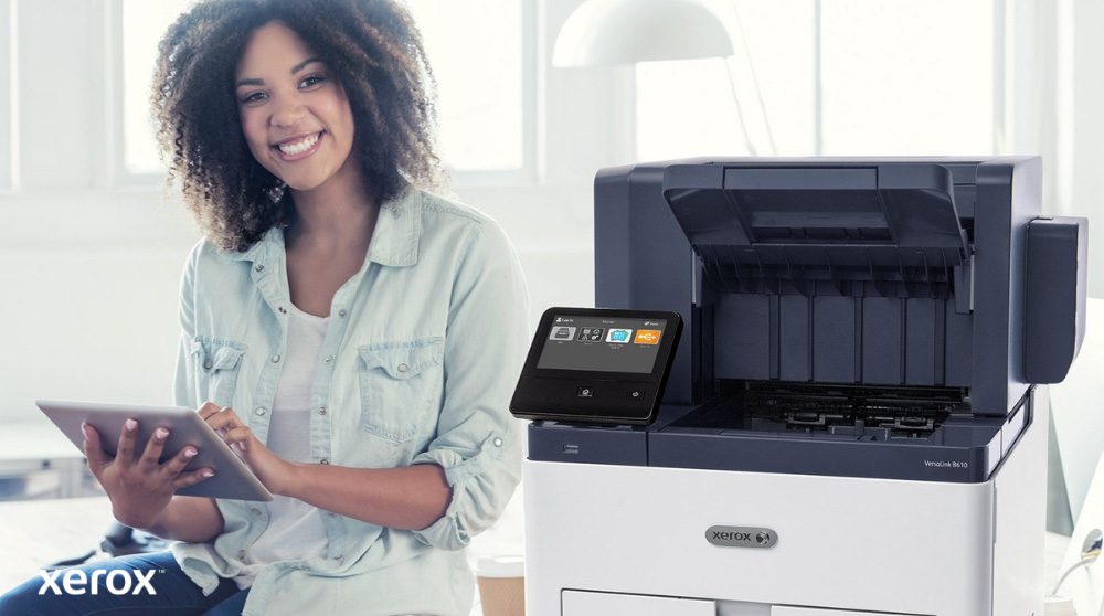 Een glimlachende persoon zit naast een witte Xerox VersaLink B610-printer, houdt een tablet vast en kijkt naar de camera. Dit benadrukt het gebruiksvriendelijke karakter van de Xerox-oplossingen.