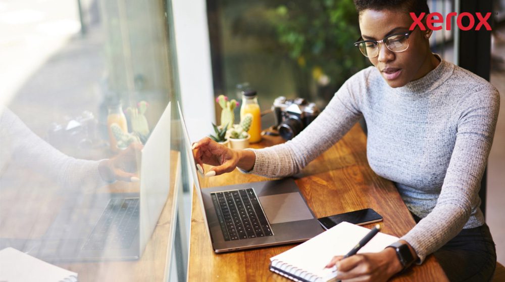 Een persoon met een bril werkt aan een houten tafel, kijkt naar een laptopscherm en schrijft in een notitieboekje, met een smartphone in de buurt, wat duidt op een productieve werkomgeving